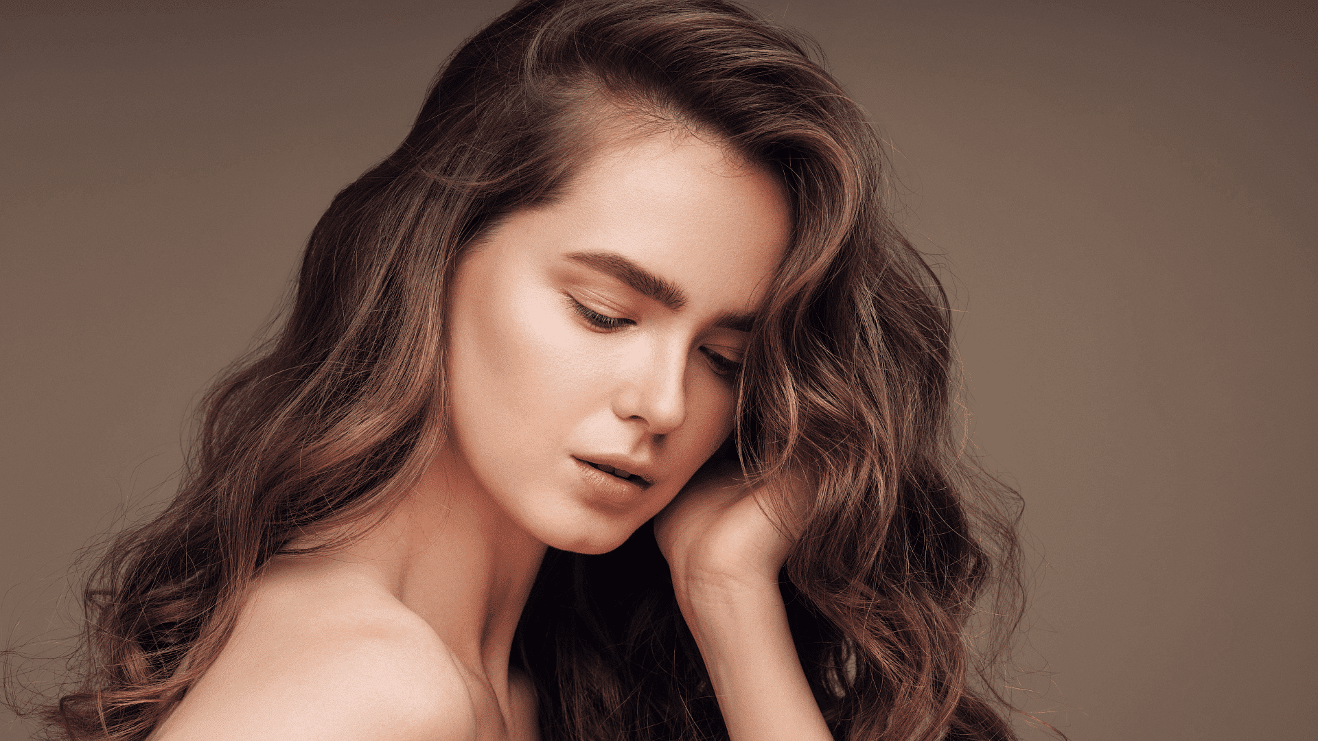 Woman with long, wavy hair looking down thoughtfully, set against a neutral background.